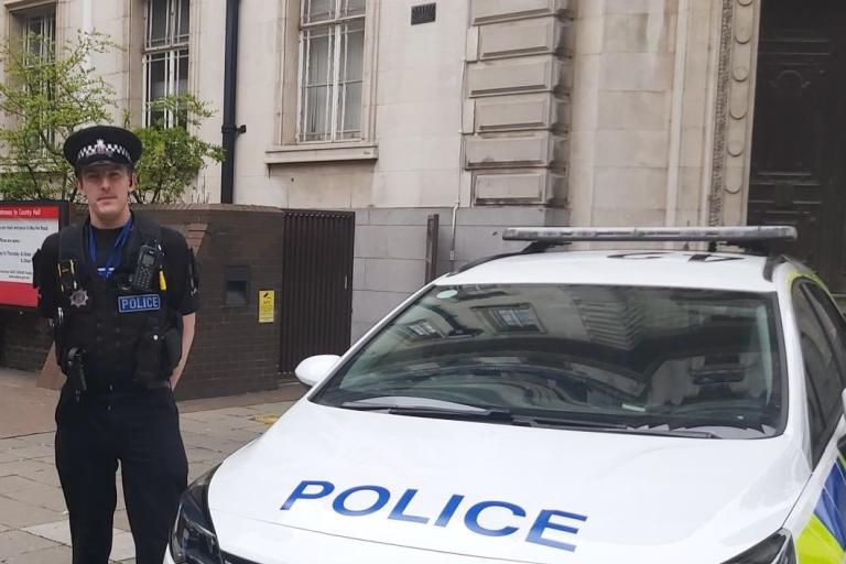 Johnathon in police uniform, next to a police car, outside County Hall