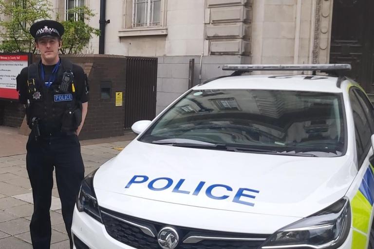 Johnathon Cuthbertson in police uniform beside a police car outside County Hall