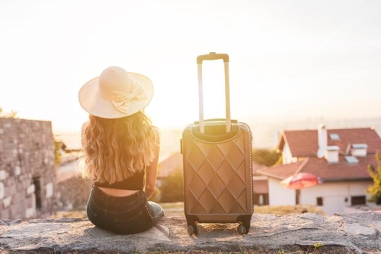 Picture of a woman sitting on a wall facing the sunset, with a suitcase beside her