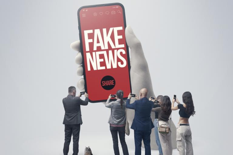 A giant mobile phone with 'fake news.share' written on it. Surrounded by a group of much smaller people taking photos of it with their mobile phones