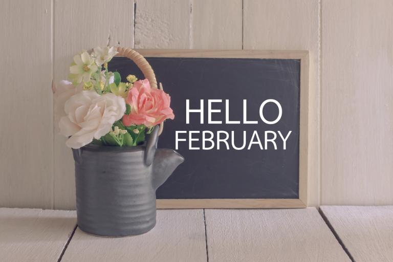 Metal watering can with white and pink roses in it, next to a chalkboard which reads Hello February