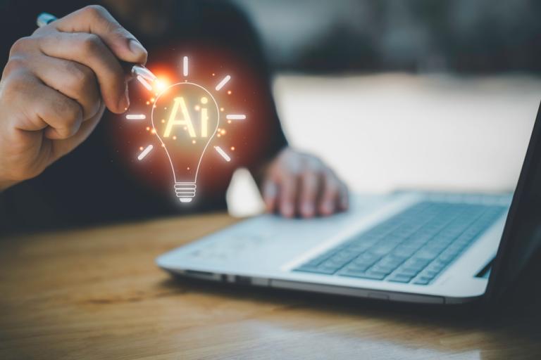 Person's hands at a laptop, with an image of a lit up light bulb floating above it with AI written inside