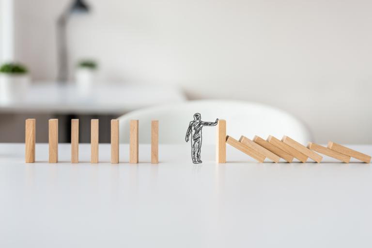Picture of a little silver stick man, in the middle of some wooden dominos. There are some on one side that have fallen down and they are holding a hand up to prevent the others being knocked over