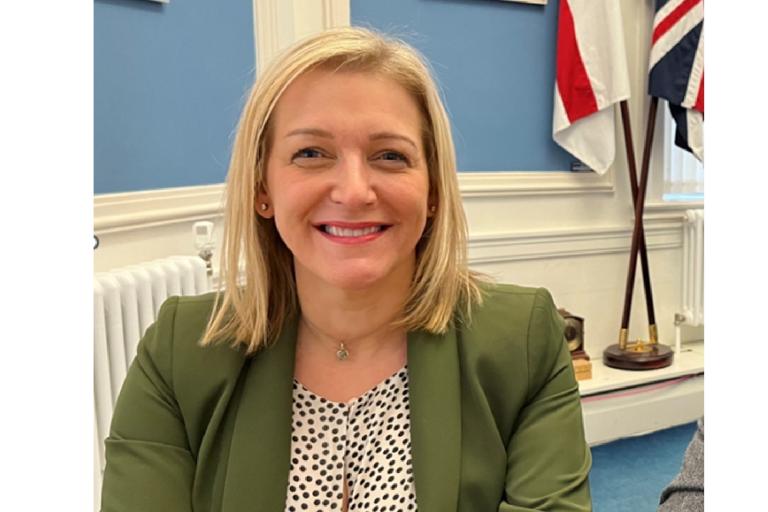 Nicole sitting smiling and wearing a green jacket. In the background and walls painted in light blue