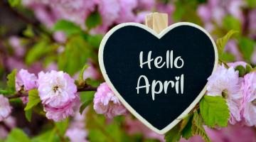 Hello April written in a heart shaped chalk board, surrounded by purple spring flowers