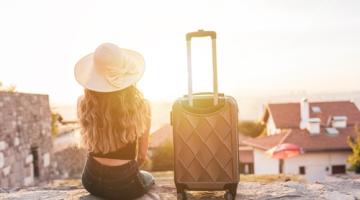 Picture of a woman sitting on a wall facing the sunset, with a suitcase beside her