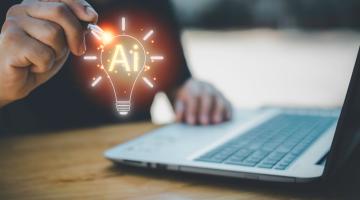 Person's hands at a laptop, with an image of a lit up light bulb floating above it with AI written inside