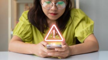 Woman wearing glasses and yellow top, holding a phone which has a hazard sign floating above it