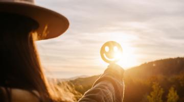 Woman in a hat standing before a setting sun, holding up a smiley face emoji
