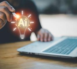 Person's hands at a laptop, with an image of a lit up light bulb floating above it with AI written inside