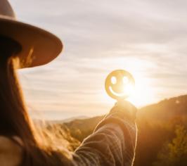 Woman in a hat standing before a setting sun, holding up a smiley face emoji